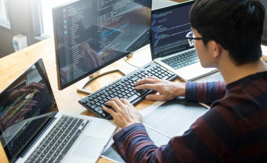 Man working on multiple monitors with code on screens in a bright office