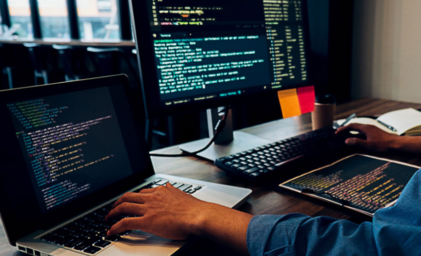 Man working on multiple monitors with code on screens in a bright office
